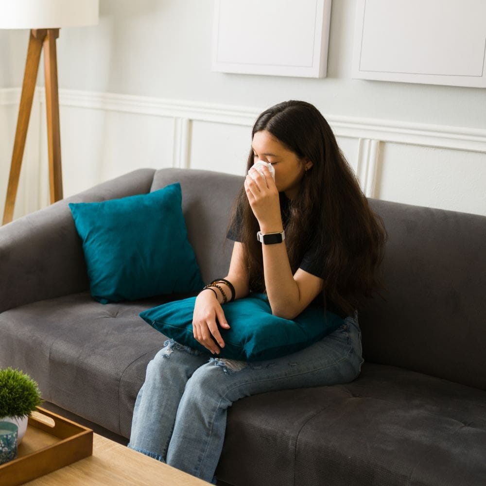 A young woman sits alone on a couch, holding a tissue and looking emotional