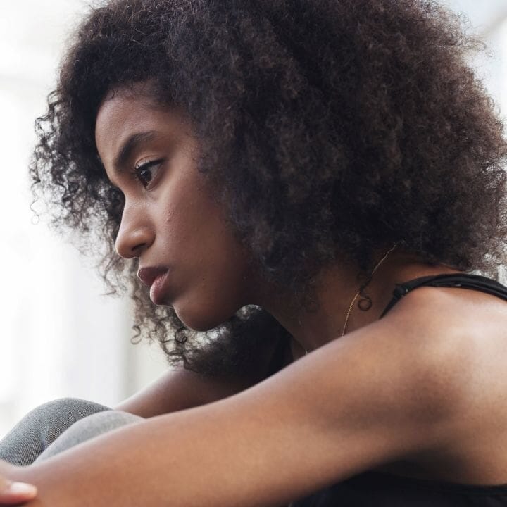 Woman sitting with knees drawn up, looking down while seated indoors.