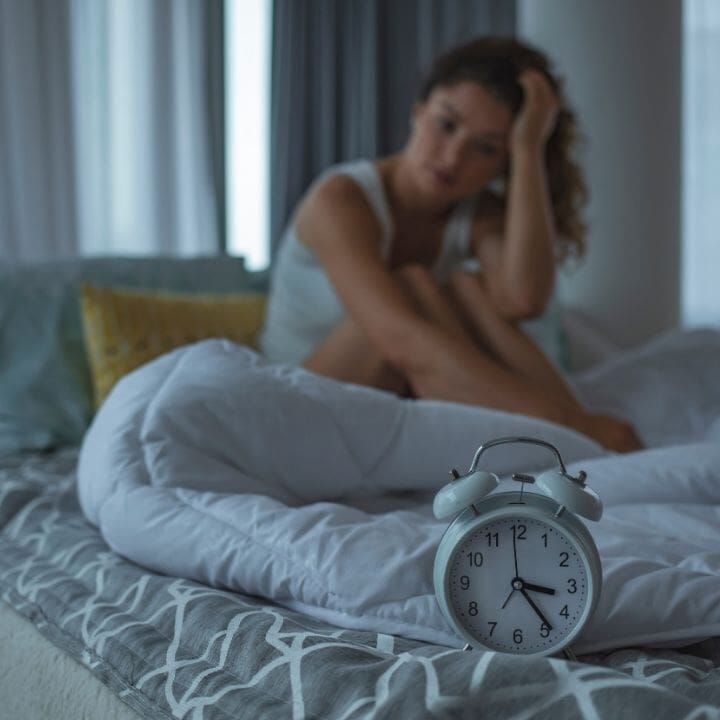 Alarm clock on bed in foreground with woman awake in background, appearing restless.
