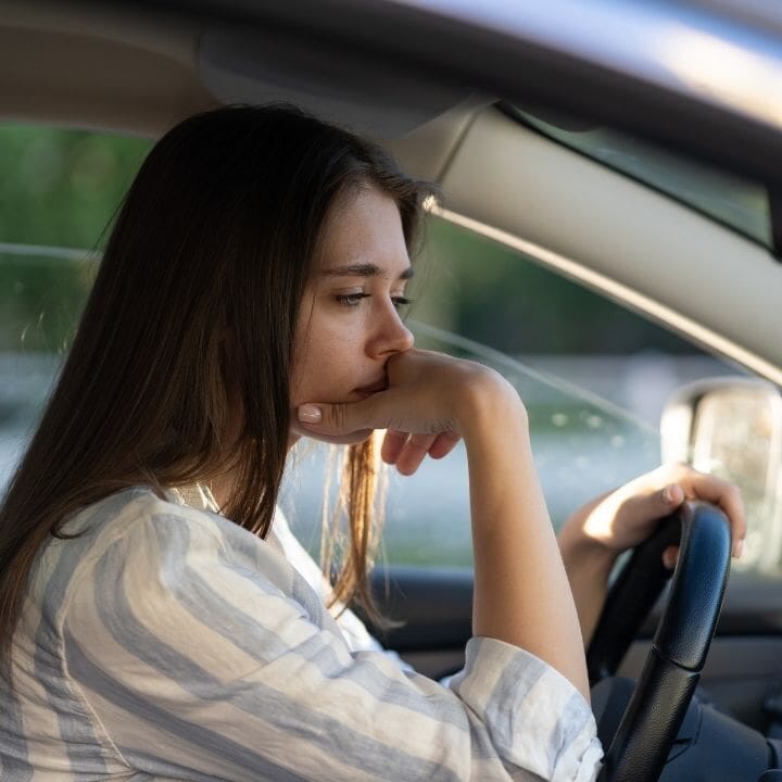 Woman seated in car holding chin, looking ahead with zoned out expression.