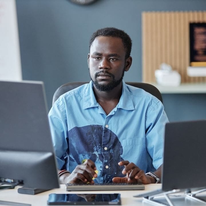 Man seated at desk between two screens with tense posture and focused expression.