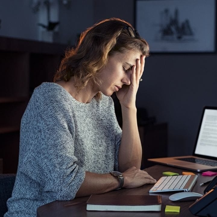 Woman at desk holding forehead beside laptop, showing signs of mental fatigue.